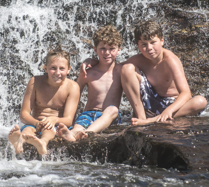 Three boys sit on rocks at a swimming hole's waterfalls.