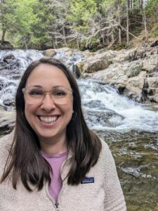 Woman with dark hair and clear glasses smiles big in front of cascades.