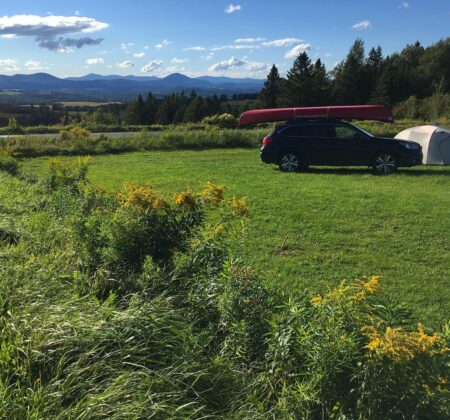 Car with canoe on top in a grassy meadow next to a tent. 