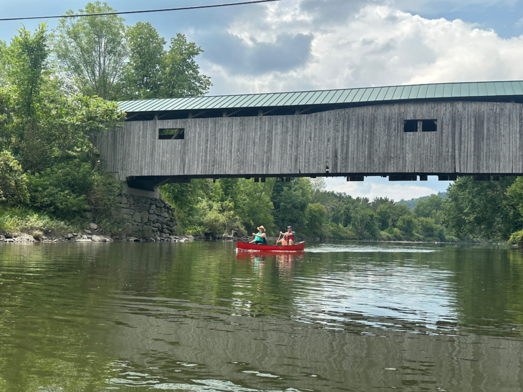 Old covered bridge crosses over the river. Two people smile, paddling a red canoe upstream toward the camera.