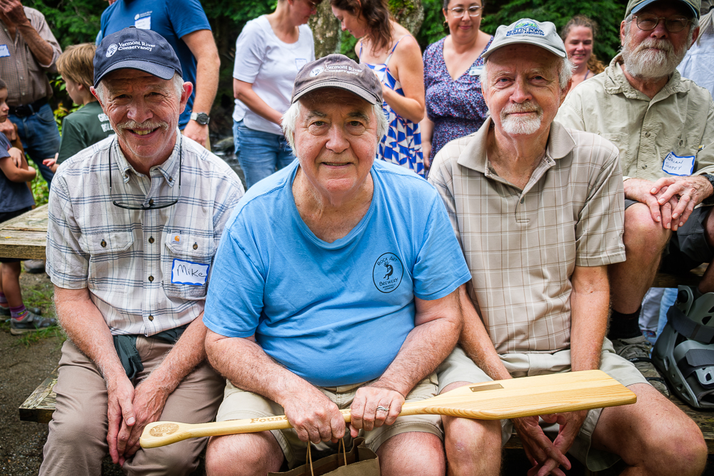 Three men sit on a bench with others in the background. All three are wearing ballcaps and smiling. The man in the middle holds a wooden paddle in his hands.